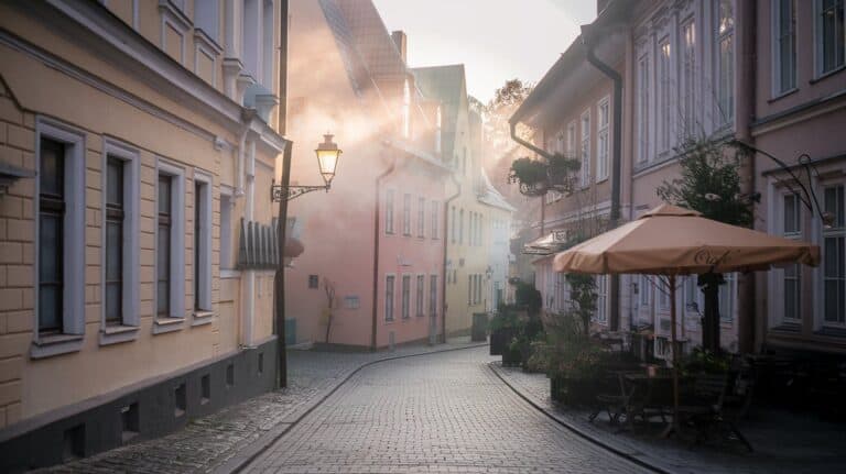 Narrow cobblestone street with pastel buildings and vintage lanterns. Morning sunlight filters through mist, casting a serene, nostalgic atmosphere.